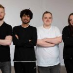 4 people stood in front of a blank wall smiling at the camera