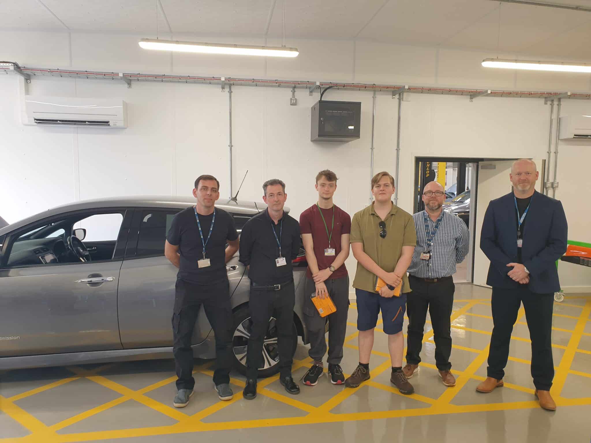 Several students and a teacher stood in a motor vehicle workshop.