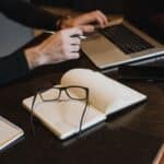 a book lying open with glasses on it and hands typing on a laptop