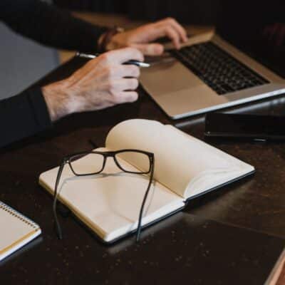 a book lying open with glasses on it and hands typing on a laptop