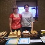 2 students stood behind a table of cakes