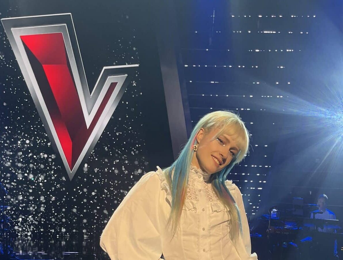 person stood on a stage in front of logo for the voice (a large red V)